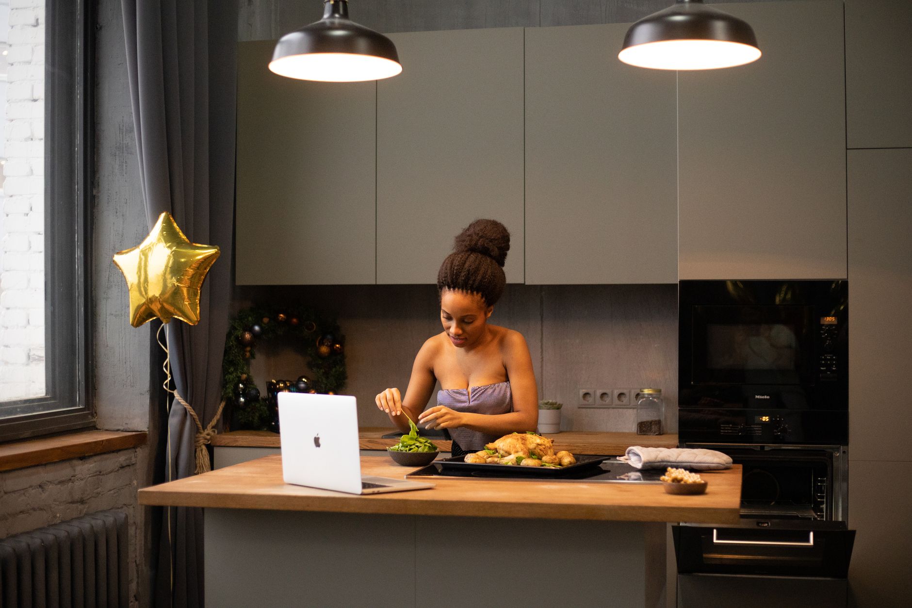 African American woman in a modern kitchen preparing food while using a laptop, under cozy lighting.